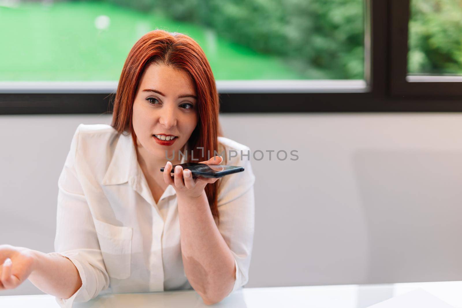 Pretty young red-haired woman looking at her smartphone while teleworking at home. Self-employed entrepreneur and businesswoman working from home or in an office while using her mobile. Large window in background with natural light.