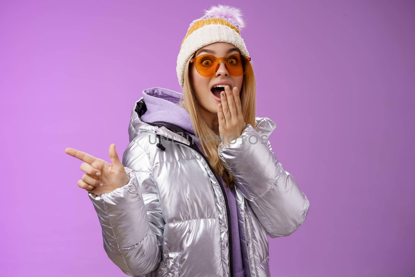 Amazed speechless surprised attractive stylish blond girlfriend in sunglasses, silver glittering jacket hat drop jaw impressed widen eyes shocked pointing left astonished, purple background by Benzoix
