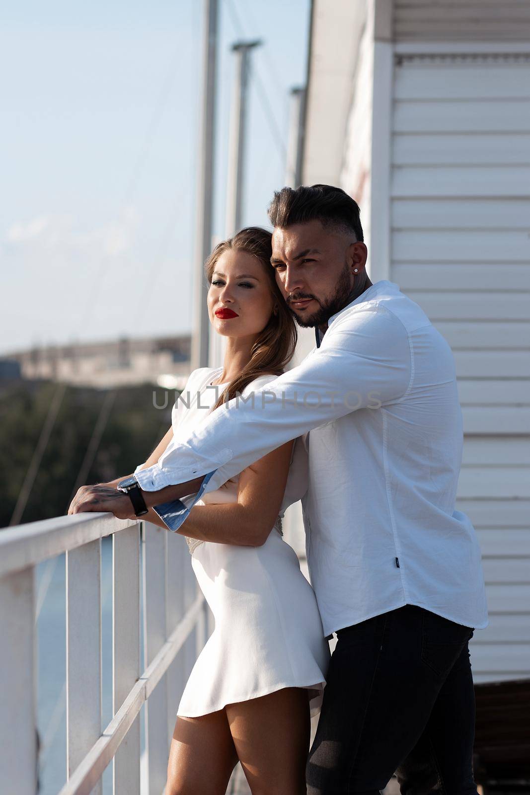 Loving couple in trendy outfits embracing while standing in port with boats in summer