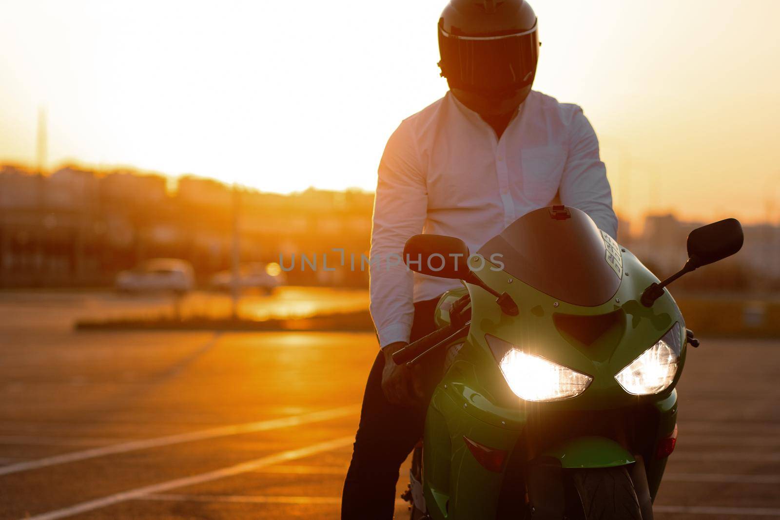 Unrecognizable stylish male motorcyclist in helmet sitting on parked motorbike in city on background of sunset sky