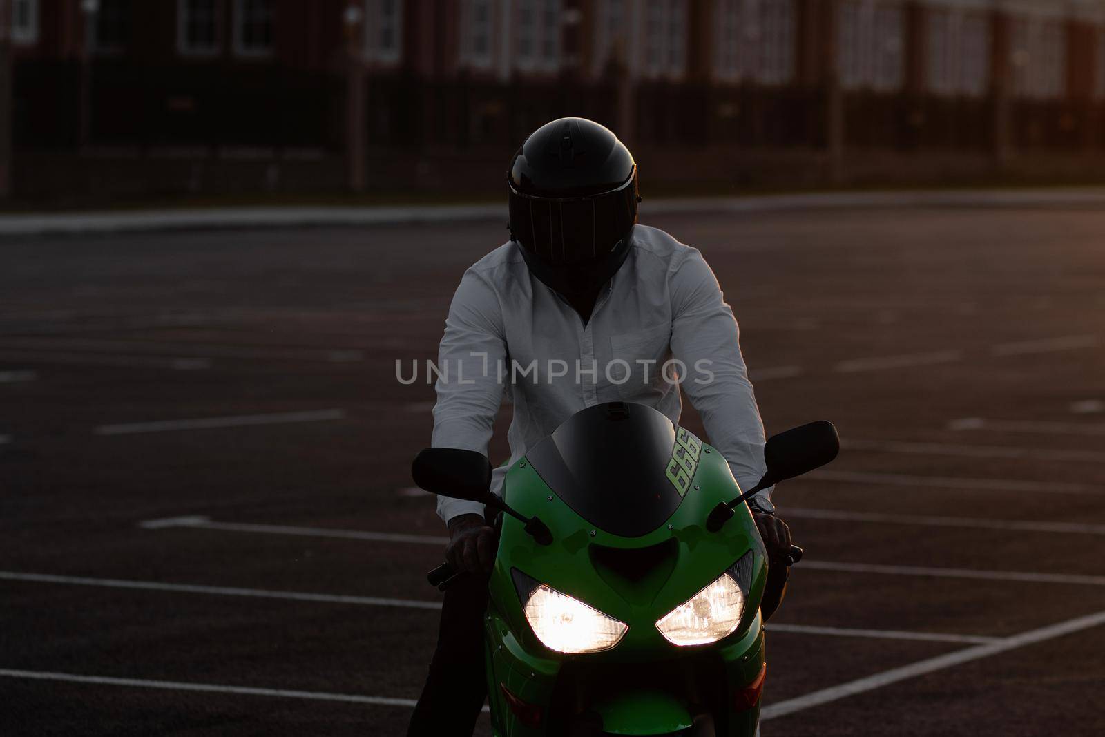 Unrecognizable stylish male motorcyclist in helmet sitting on parked motorbike in city on background of sunset sky