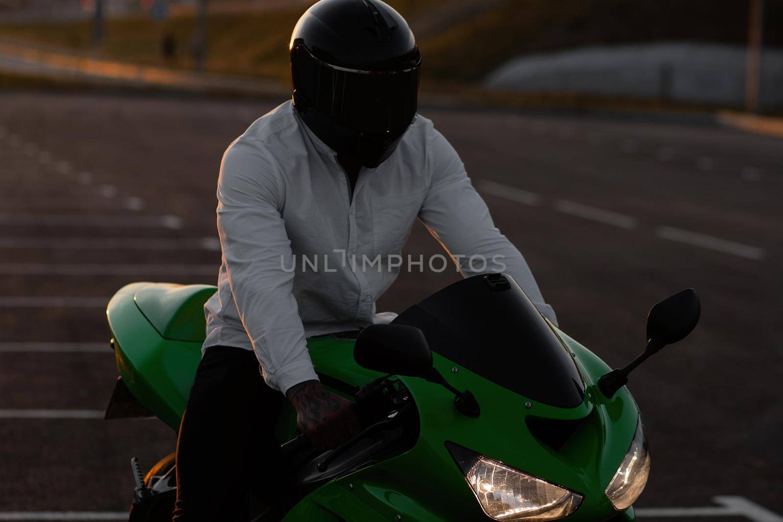 Unrecognizable stylish male motorcyclist in helmet sitting on parked motorbike in city on background of sunset sky