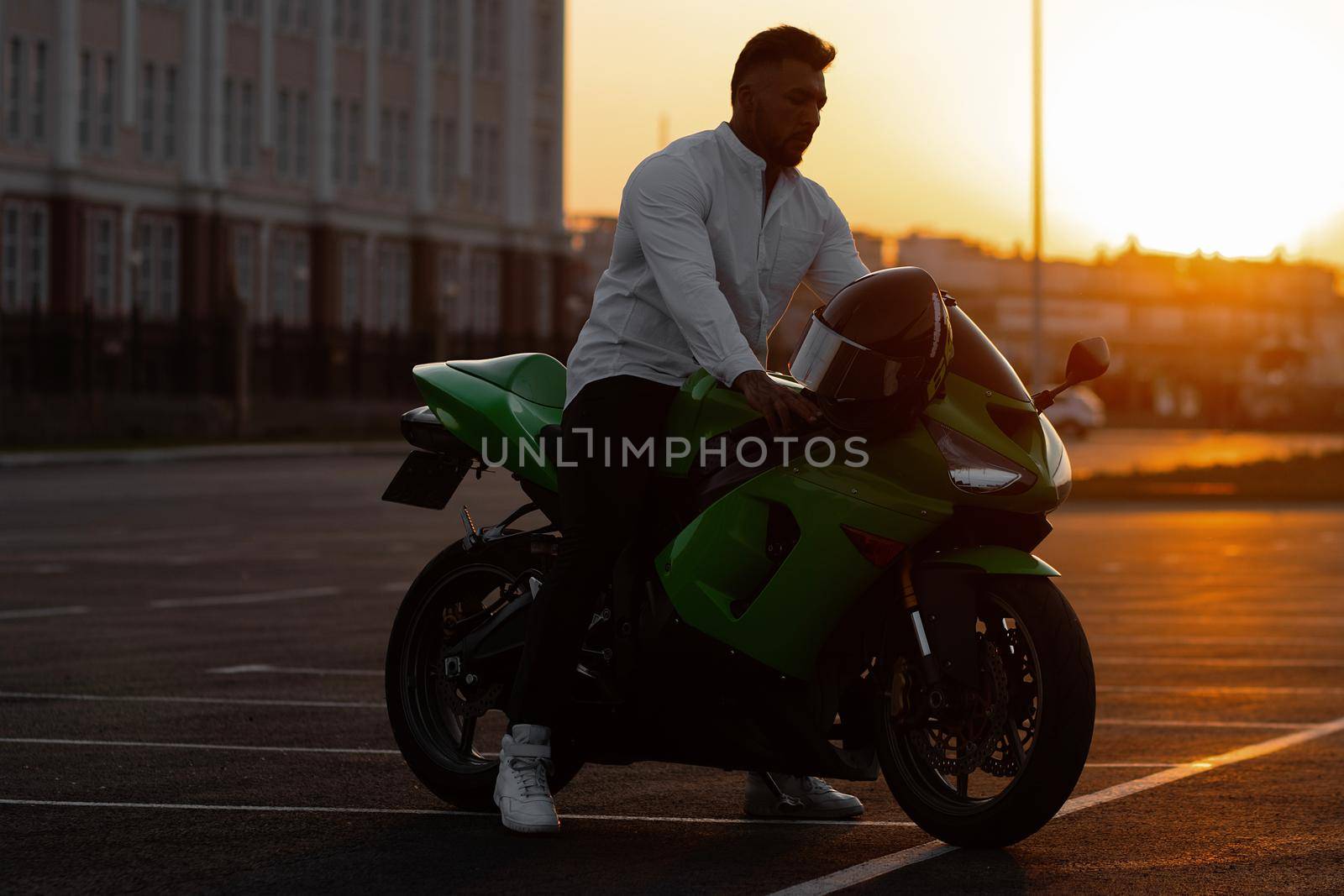 Side view of handsome male motorcyclist sitting on modern cool motorbike parked on asphalt parking against sunset sky