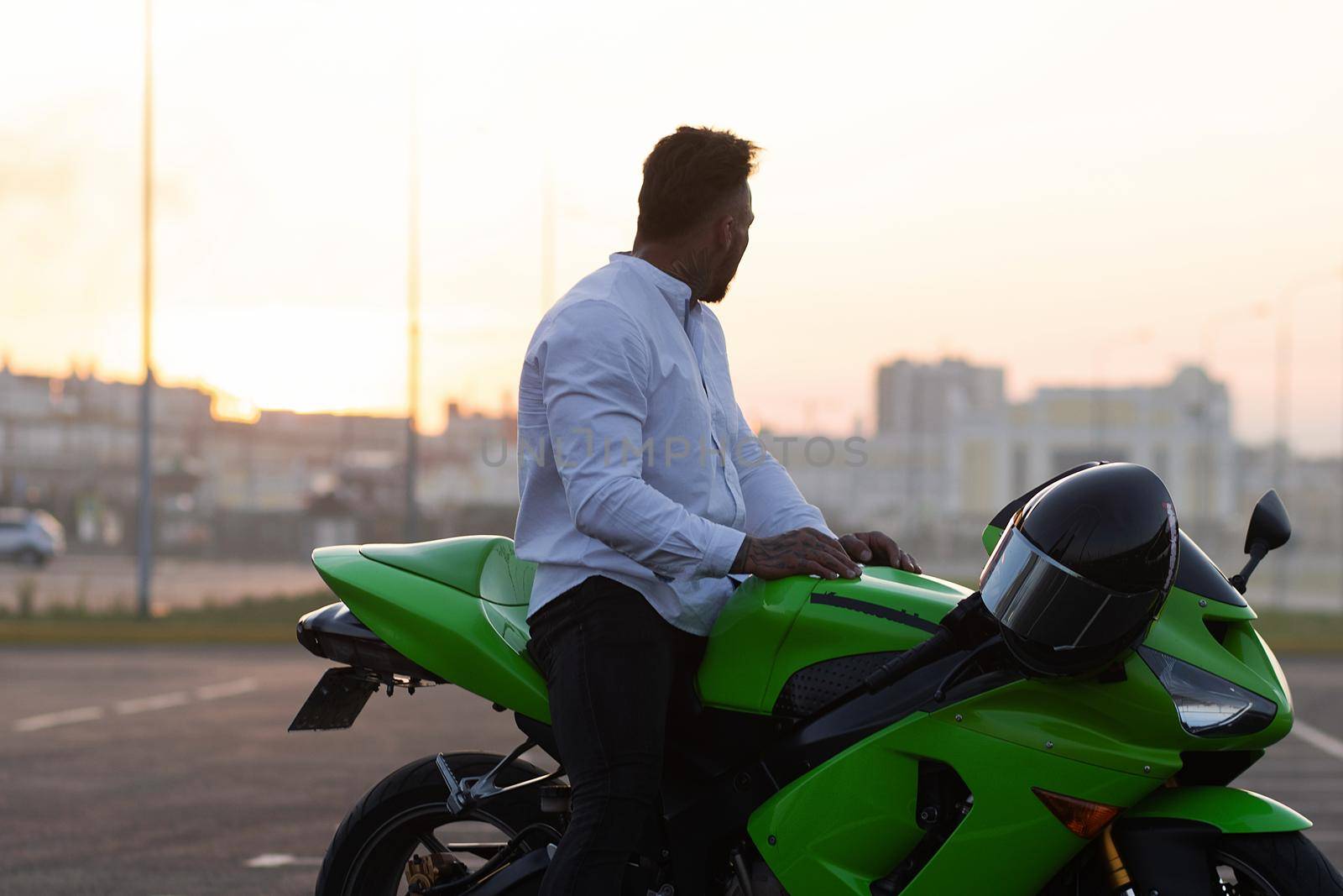 Side view of handsome male motorcyclist sitting on modern cool motorbike parked on asphalt parking against sunset sky