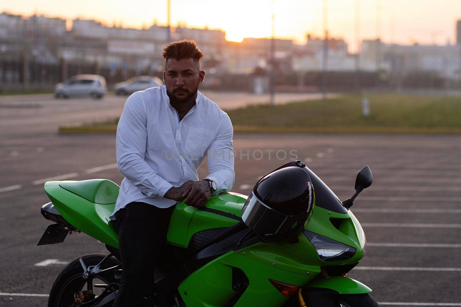 Side view of handsome male motorcyclist sitting on modern cool motorbike parked on asphalt parking against sunset sky