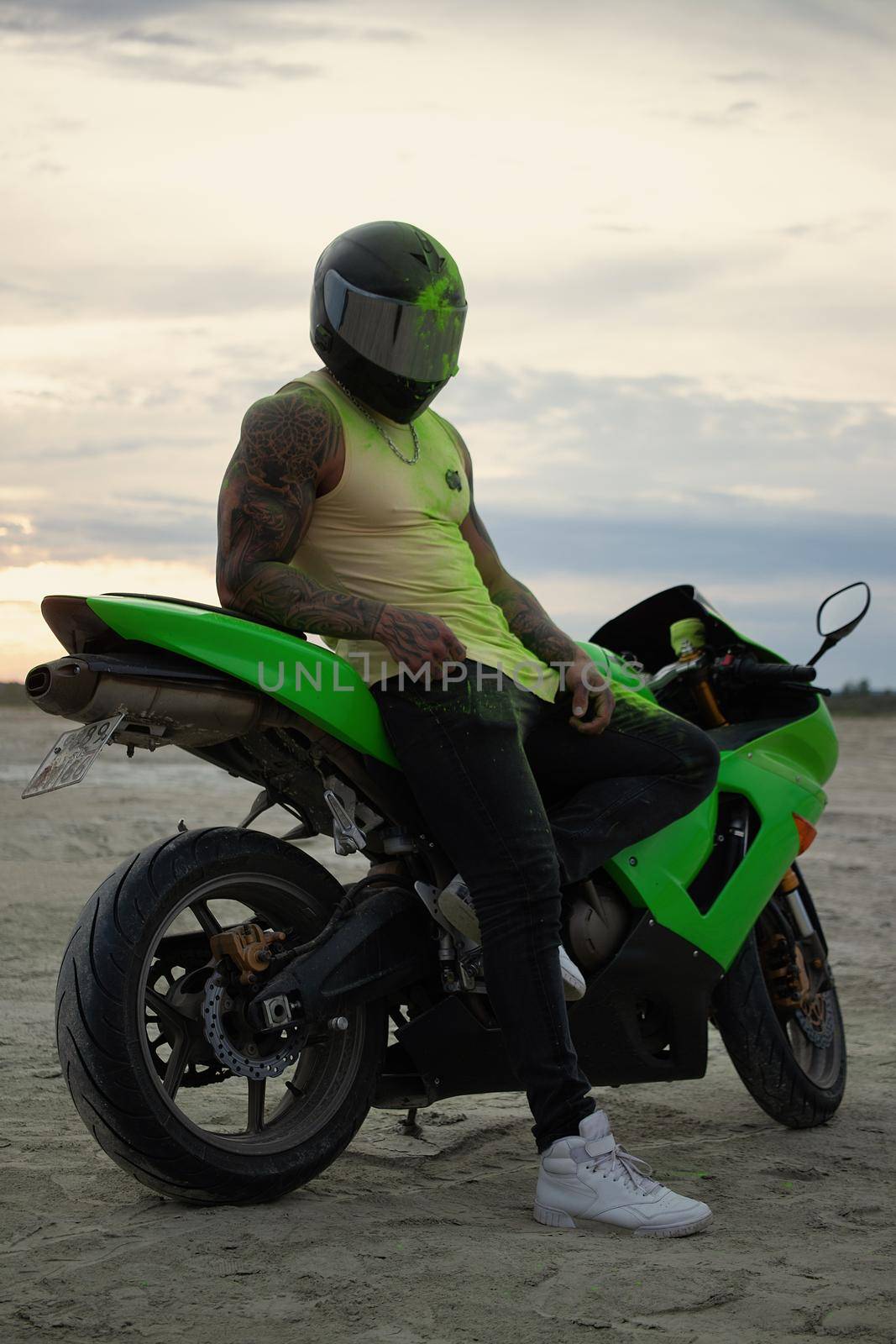 Side view of stylish male motorcyclist in helmet standing near modern motorbike on sandy seashore in evening