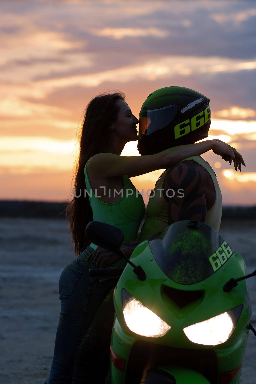 Side view of couple in love tenderly hugging near motorcycle parked on sandy seashore against sundown sky
