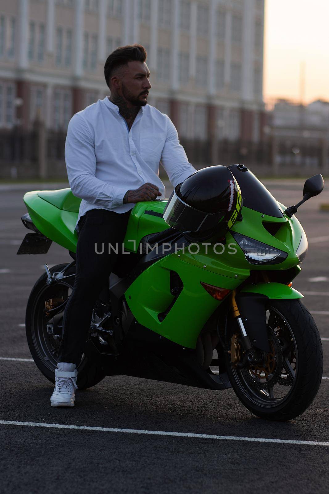 Side view of handsome male motorcyclist sitting on modern cool motorbike parked on asphalt parking against sunset sky