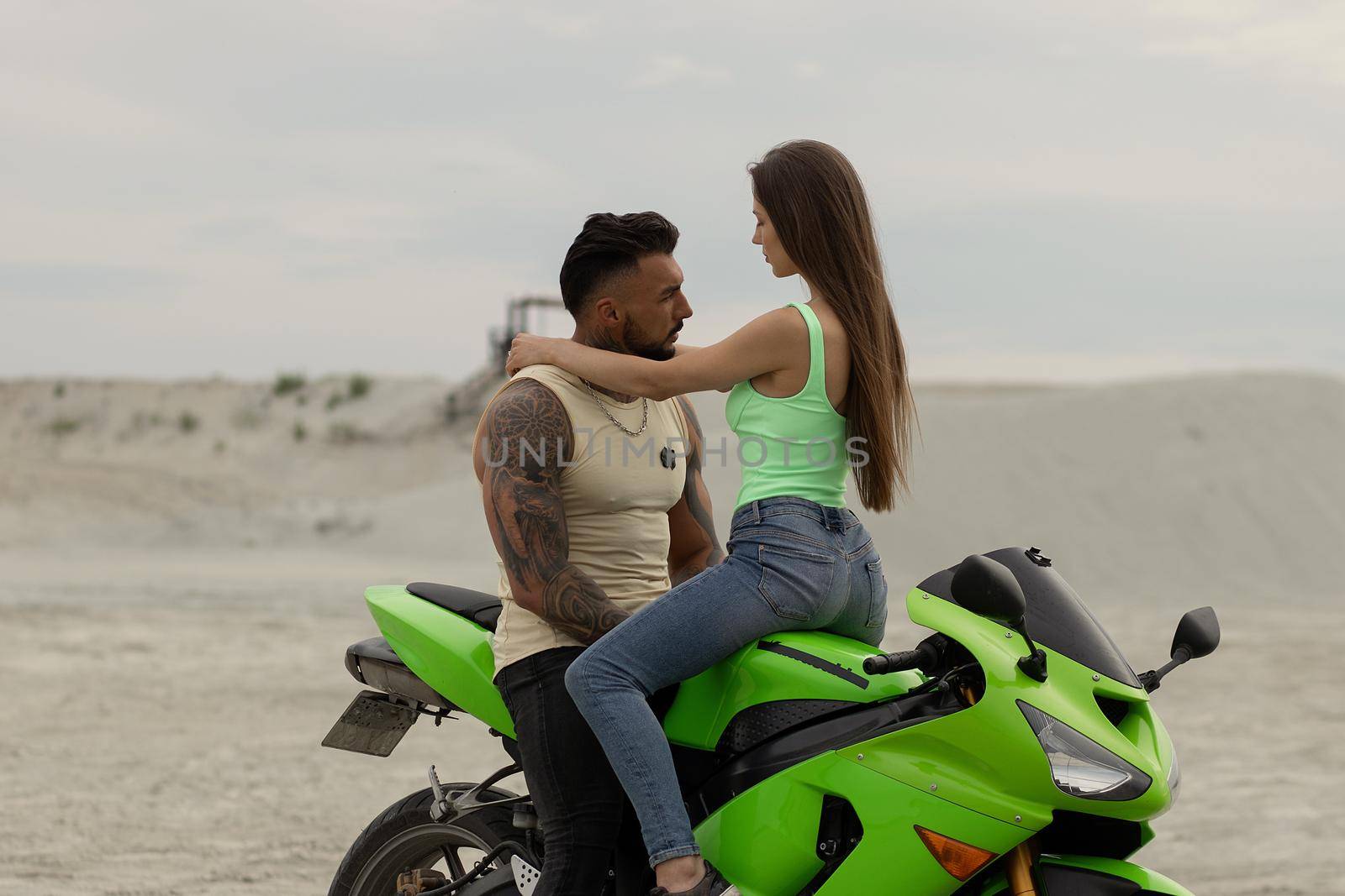 Young cool couple sitting on modern motorcycle parked on sandy beach in summer evening
