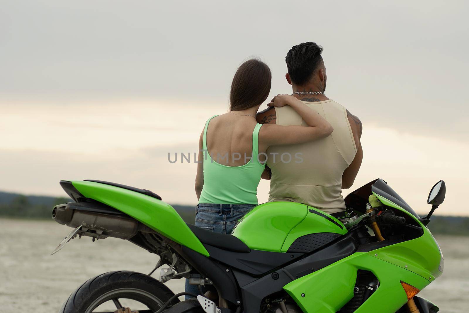 Young cool couple sitting on modern motorcycle parked on sandy beach in summer evening