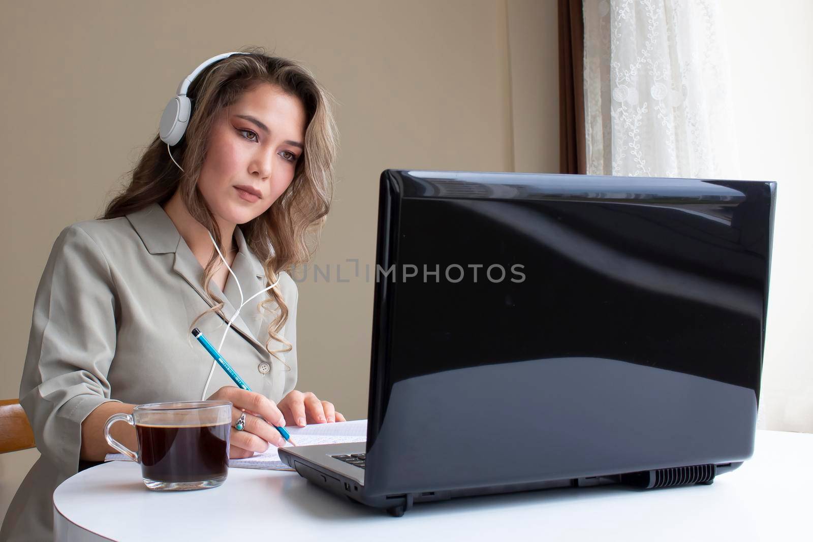 beautiful asian girl is sitting with headphone at a computer to study or work. there are a cup of coffee, a copybook and a pencil on a table