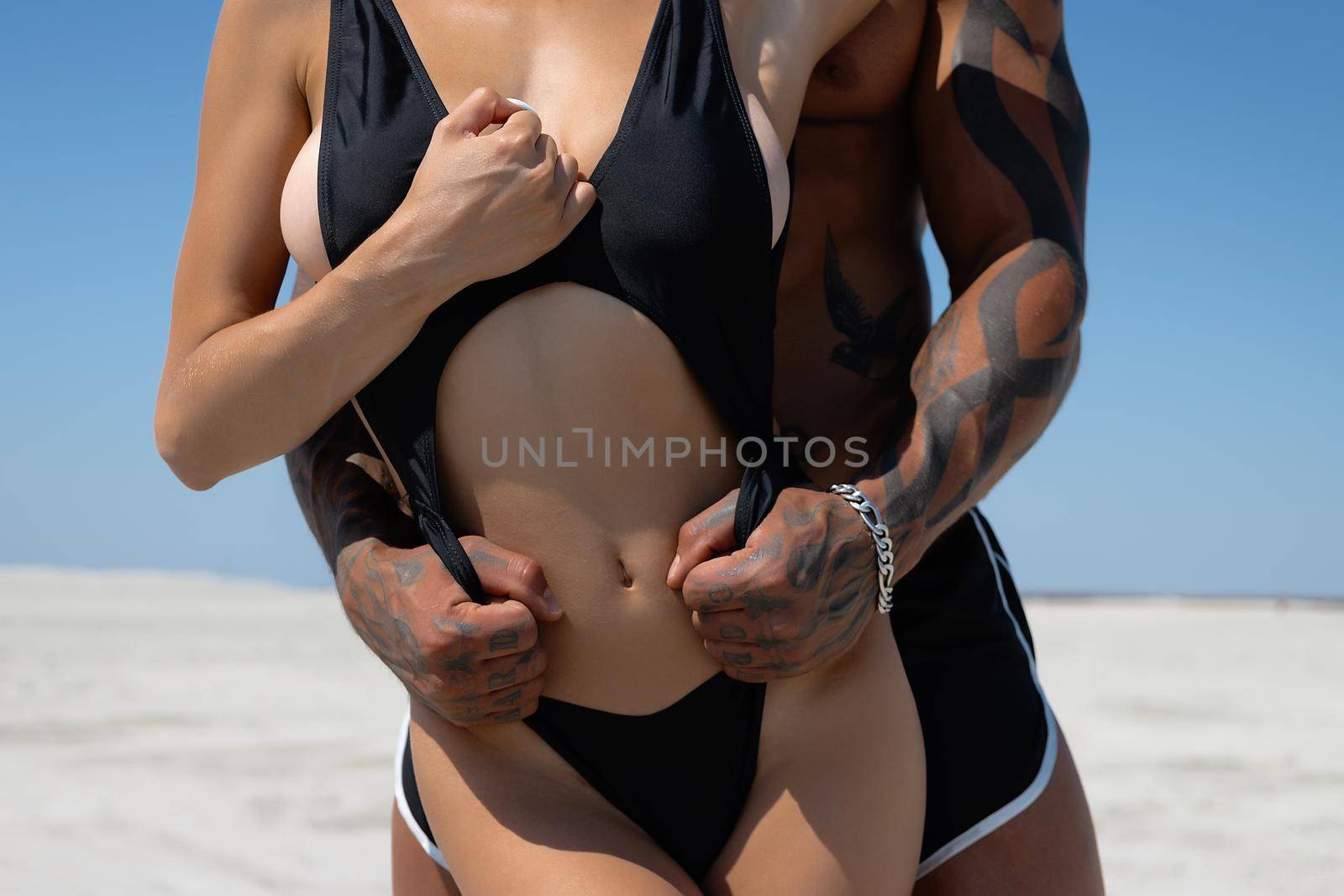 Seductive female in swimsuit and fit male with naked torso hugging while standing on sandy seashore on sunny day in summer