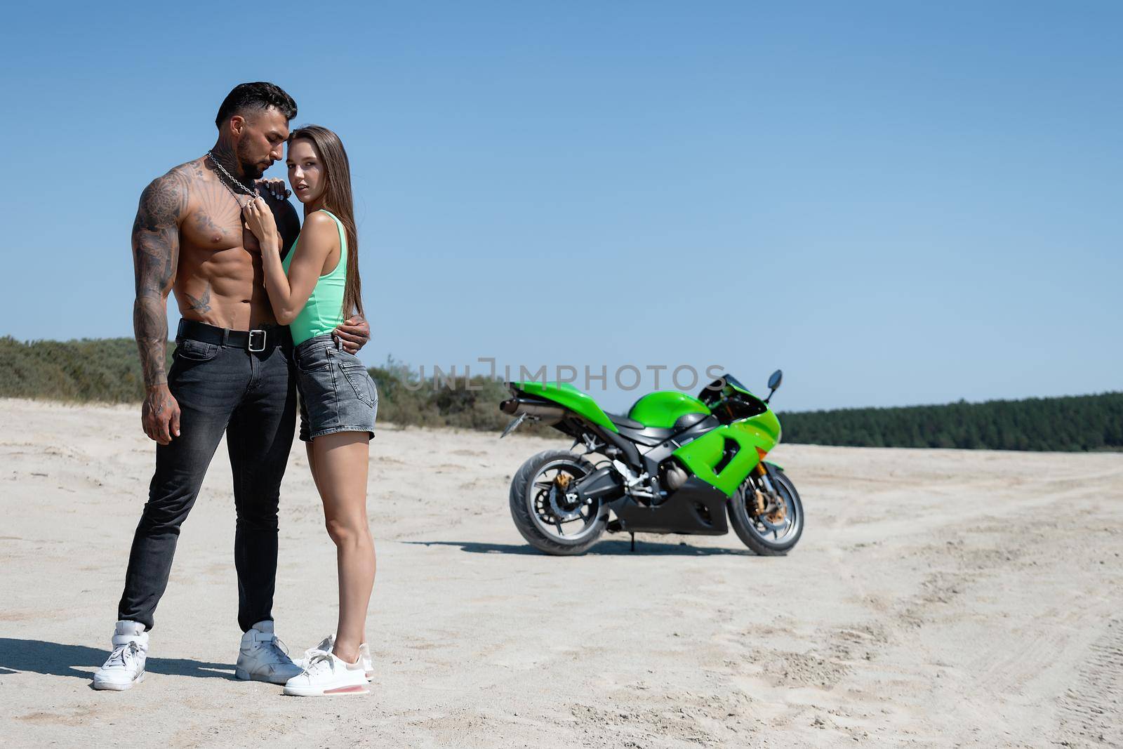 Serious male with tattoos and beard standing and embracing sensual girlfriend with long brown hair against green motorbike in sandy field in nature in sunny day