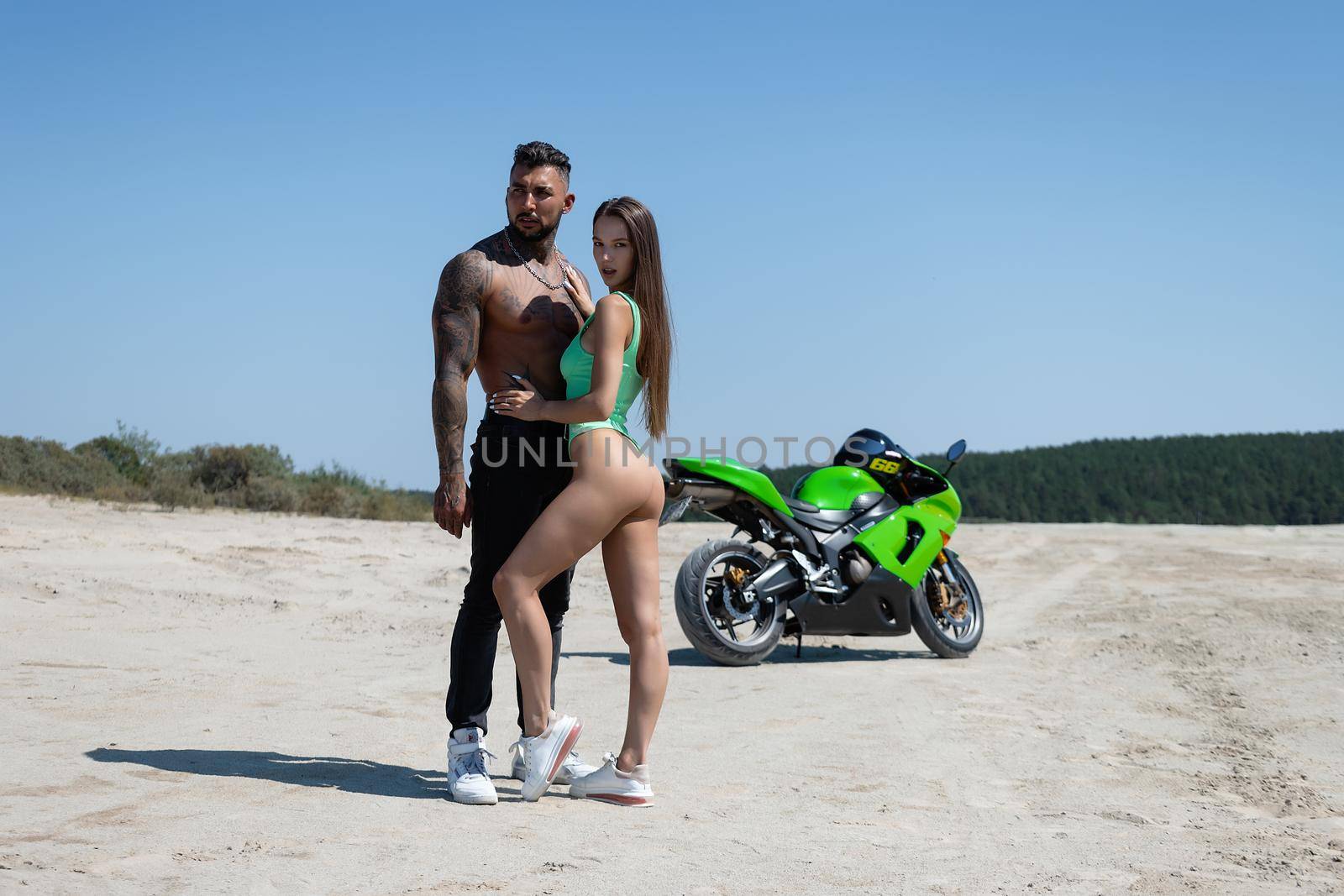 Serious male with tattoos and beard standing and embracing sensual girlfriend with long brown hair against green motorbike in sandy field in nature in sunny day