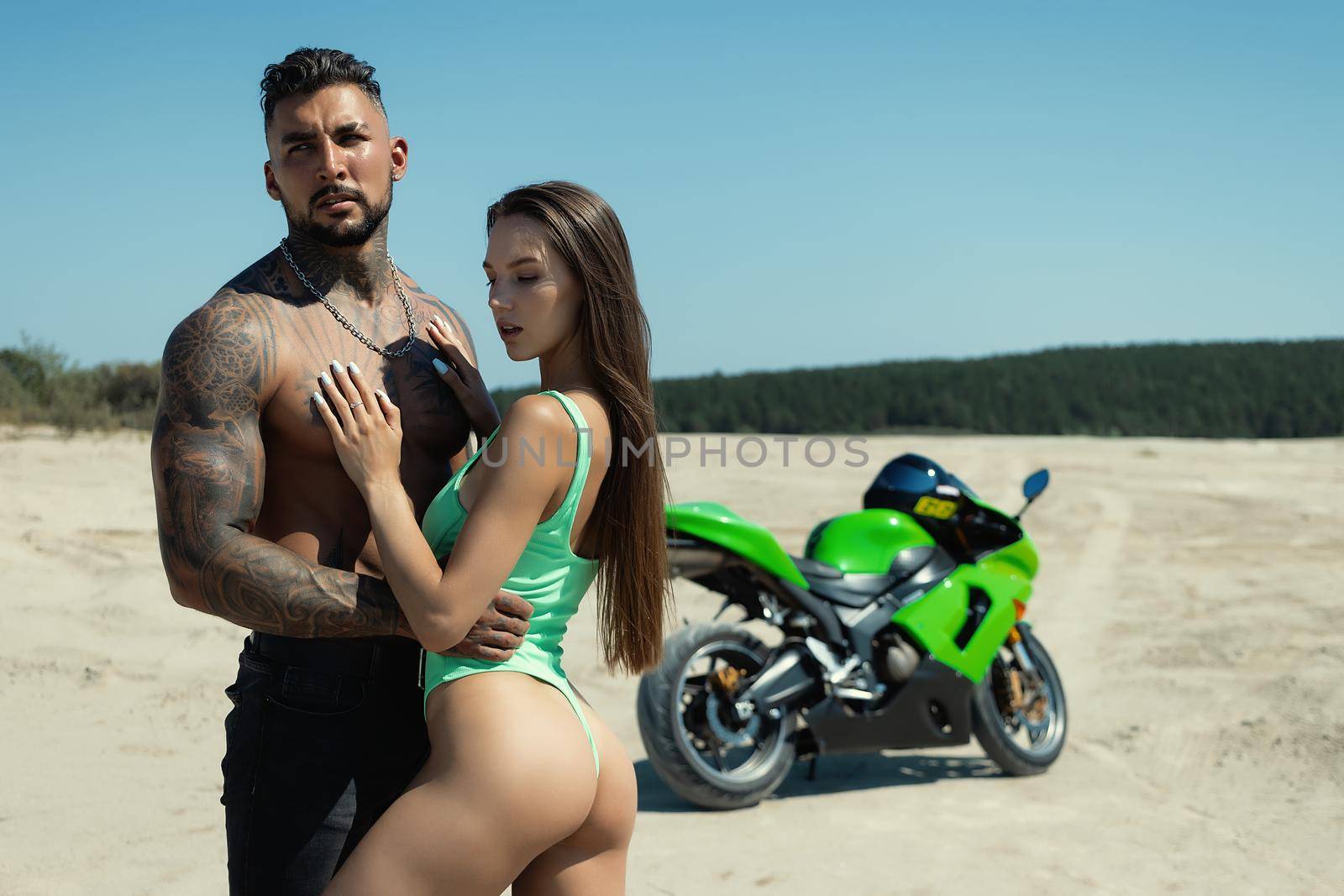Serious male with tattoos and beard standing and embracing sensual girlfriend with long brown hair against green motorbike in sandy field in nature in sunny day
