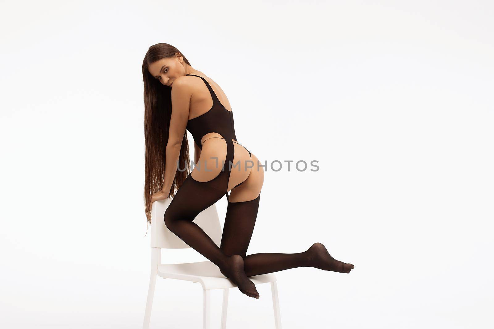 Full body of sexual unrecognizable female model in black lingerie with stockings leaning on chair against white background in light studio