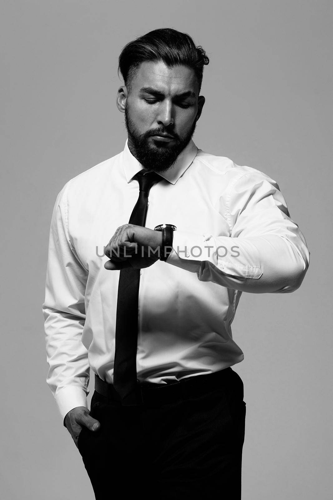 Bearded Hispanic guy in dark vest and white shirt with tie looking at camera with hands in pockets in studio