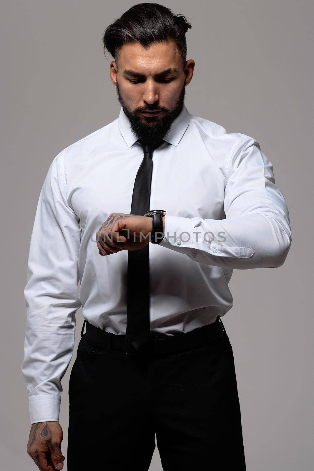 Bearded Hispanic guy in dark vest and white shirt with tie looking at camera with hands in pockets in studio