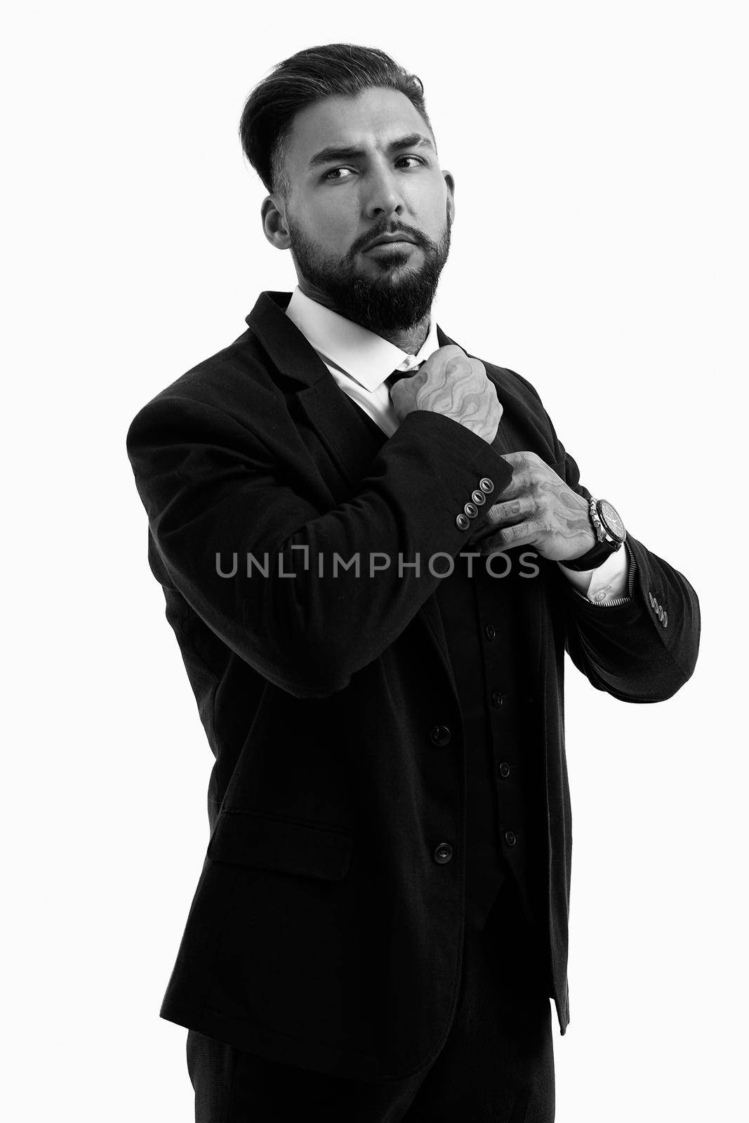 Confident Hispanic bearded man with tattooed hands in white shirt and black trousers adjusting tie and looking away while standing against gray background