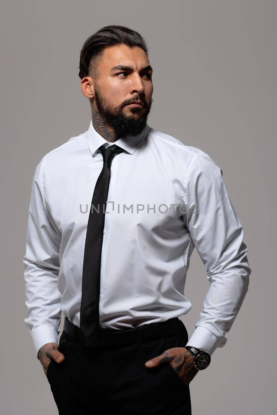 Bearded Hispanic guy in dark vest and white shirt with tie looking at camera with hands in pockets in studio