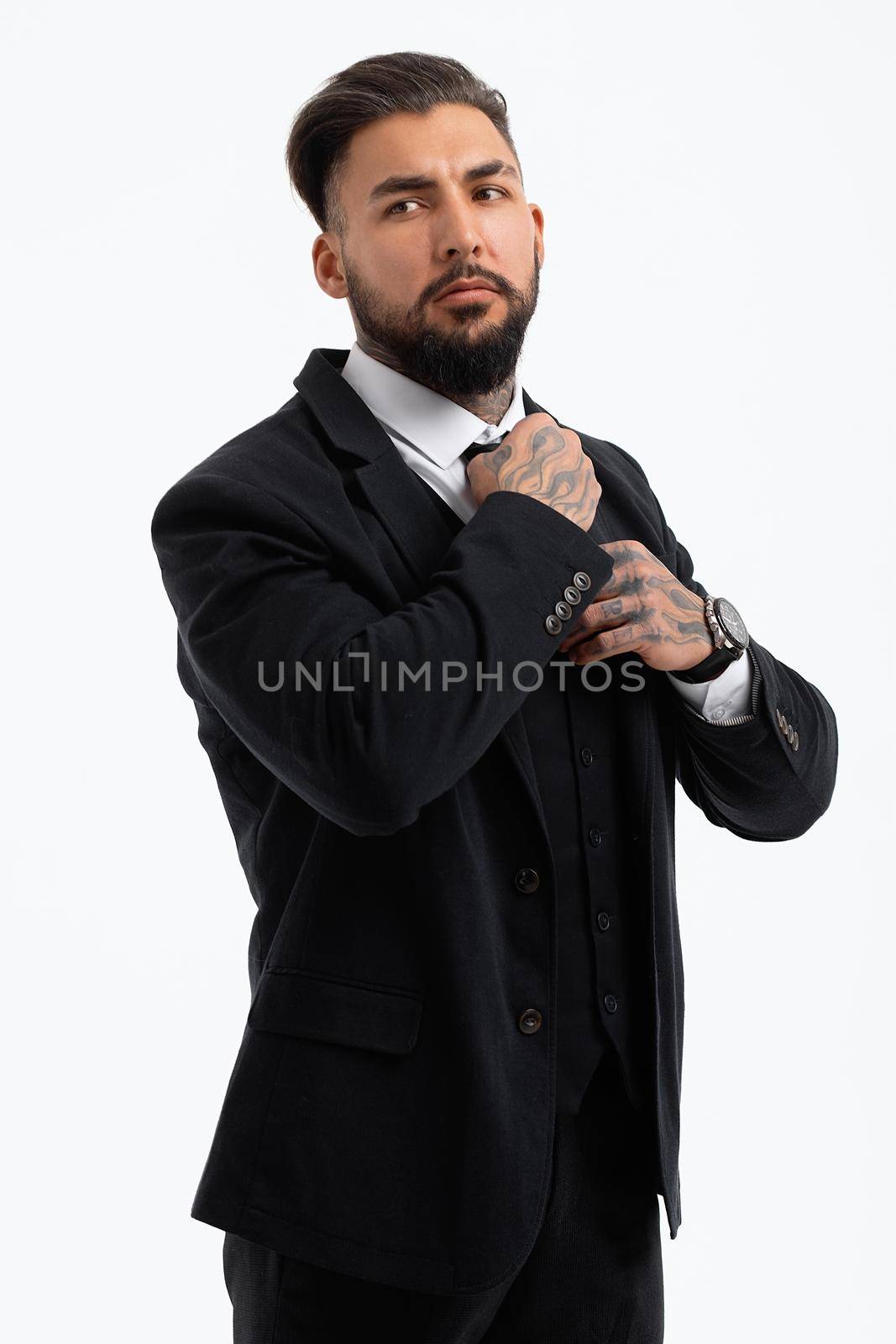 Confident Hispanic bearded man with tattooed hands in white shirt and black trousers adjusting tie and looking away while standing against gray background