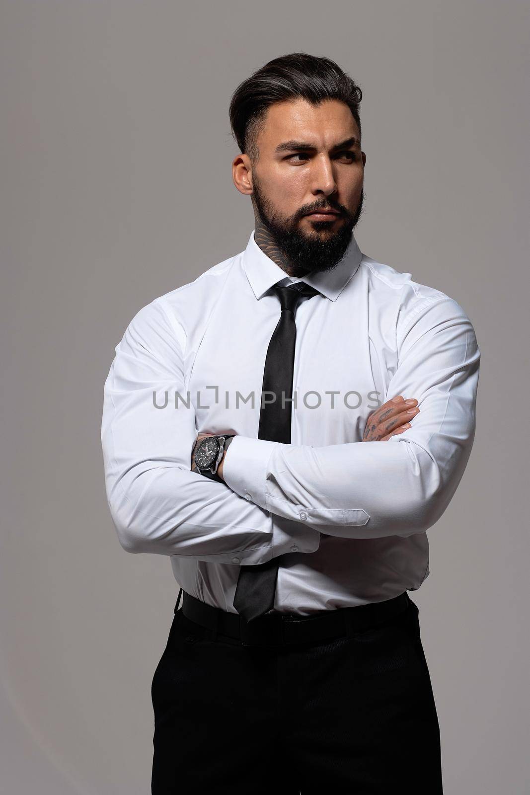 Bearded Hispanic guy in dark vest and white shirt with tie looking at camera with hands in pockets in studio