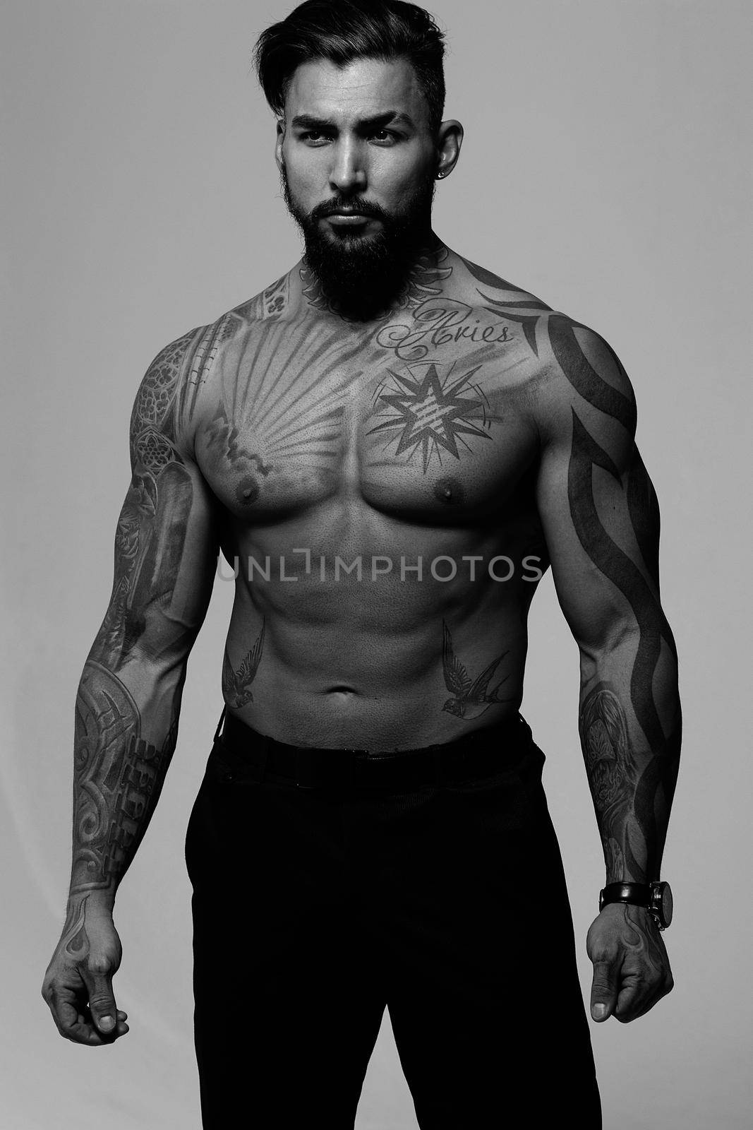 Hispanic shirtless male model with muscular tattooed torso standing with hands in pockets and looking away on gray backdrop