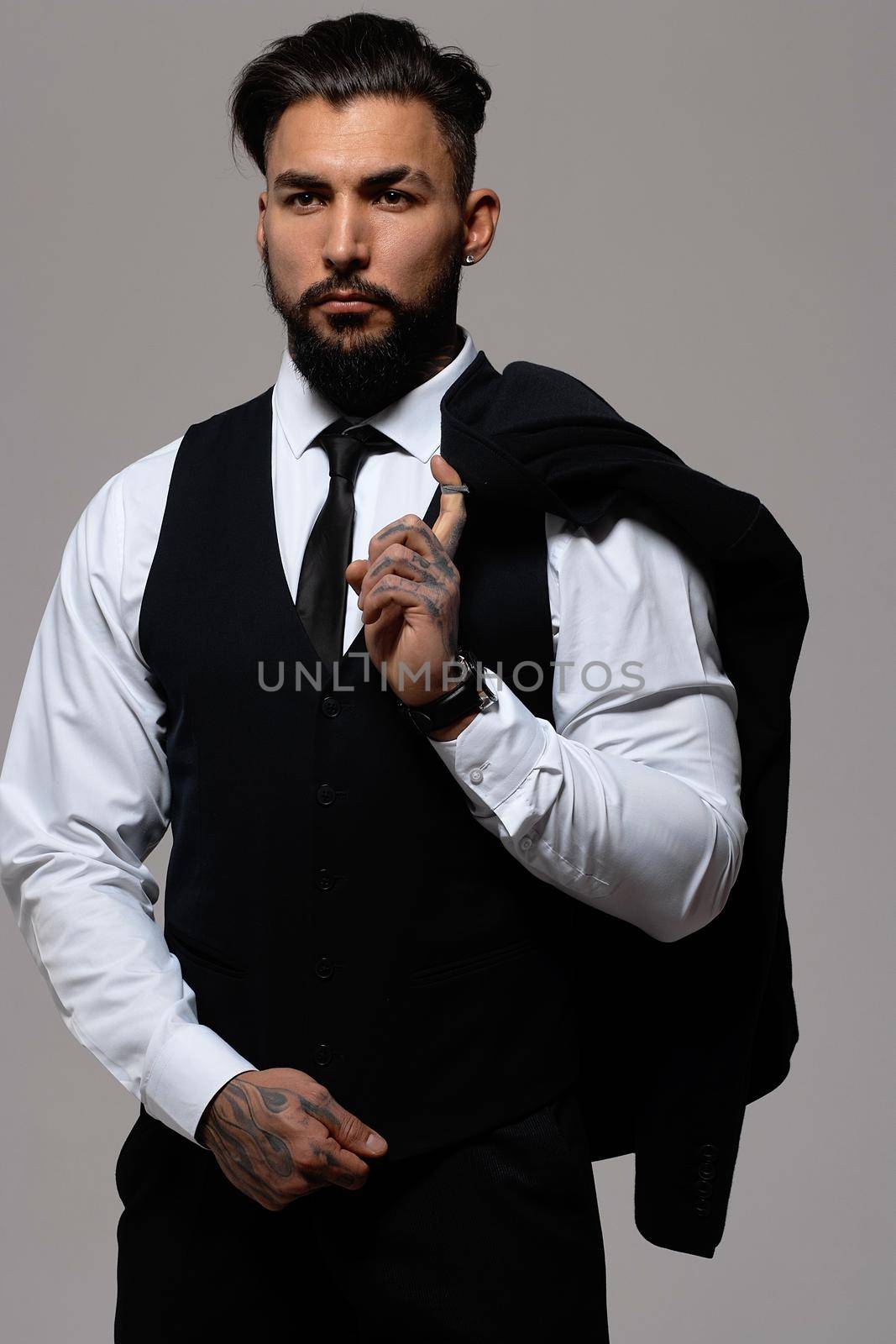 Bearded Hispanic guy in dark vest and white shirt with tie looking at camera with hands in pockets in studio