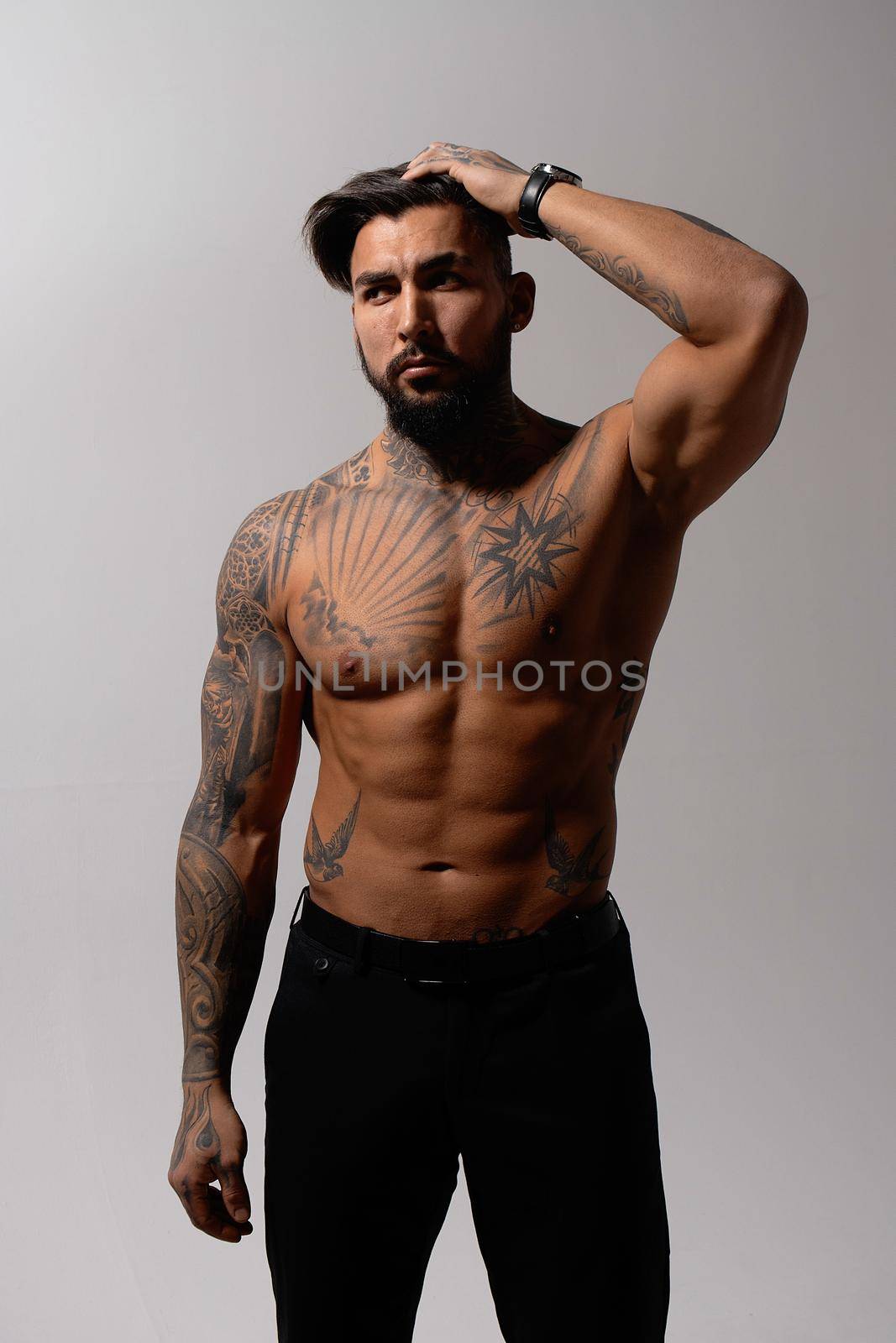 Hispanic shirtless male model with muscular tattooed torso standing with hands in pockets and looking away on gray backdrop