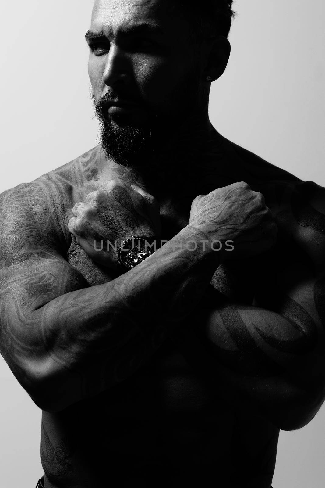 Hispanic shirtless male model with muscular tattooed torso standing with hands in pockets and looking away on gray backdrop