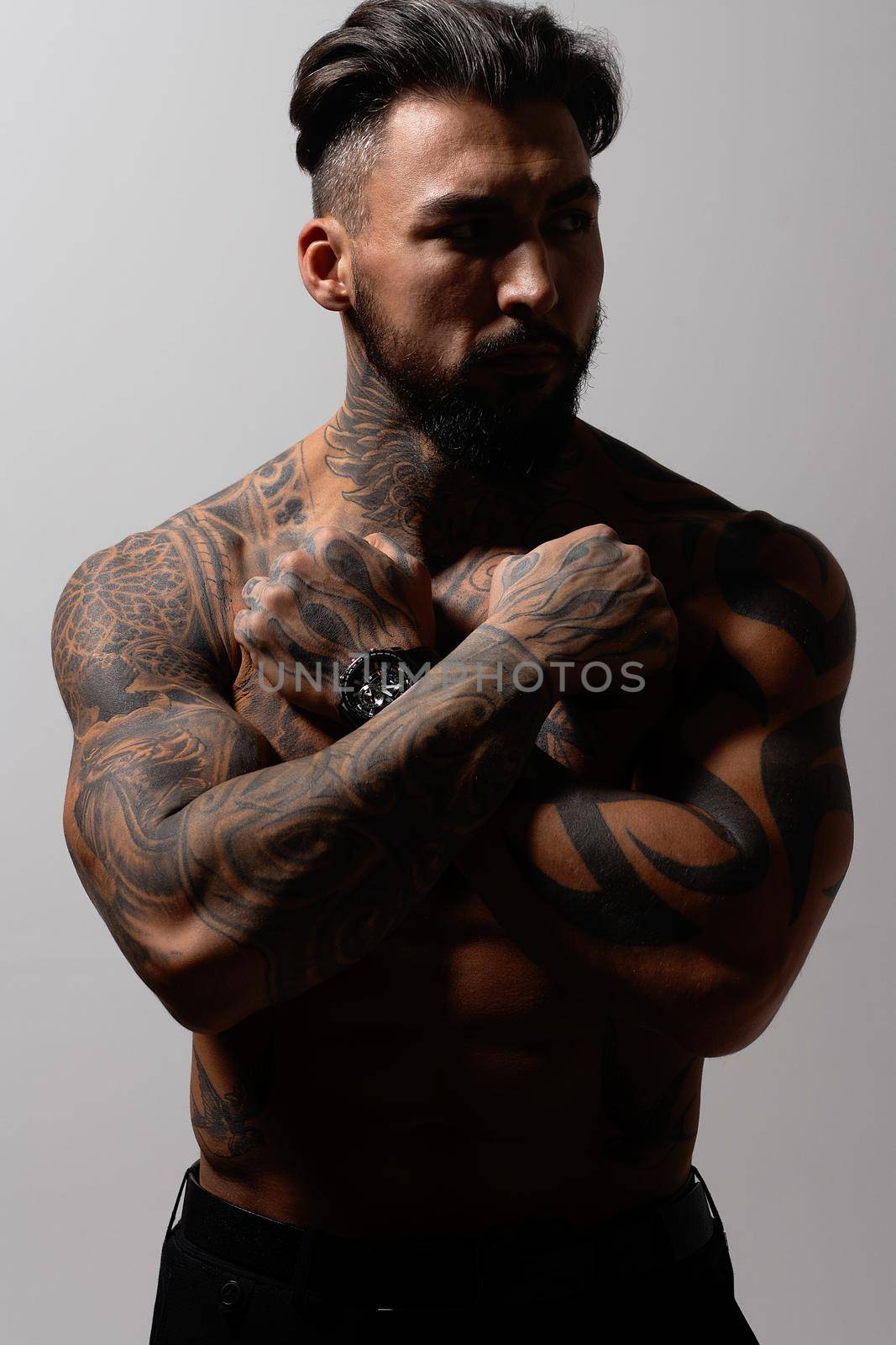 Hispanic shirtless male model with muscular tattooed torso standing with hands in pockets and looking away on gray backdrop