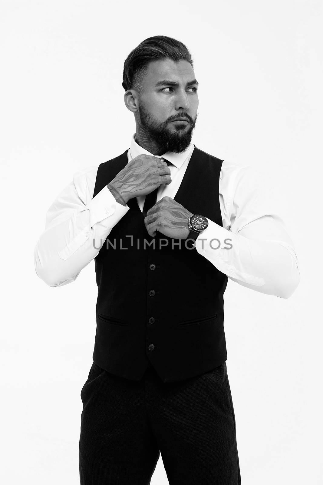 Confident Hispanic bearded man with tattooed hands in white shirt and black trousers adjusting tie and looking away while standing against gray background