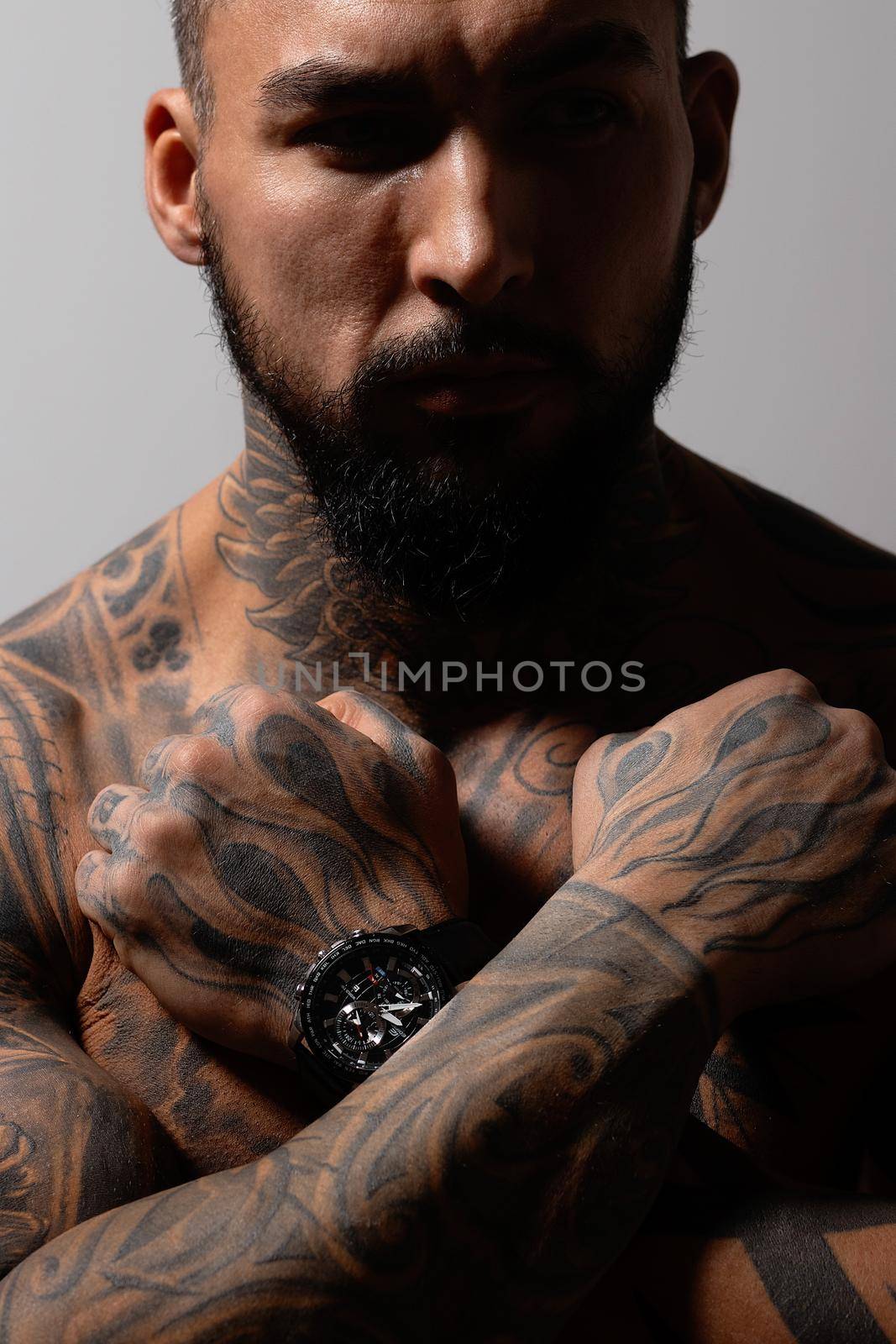 Hispanic shirtless male model with muscular tattooed torso standing with hands in pockets and looking away on gray backdrop