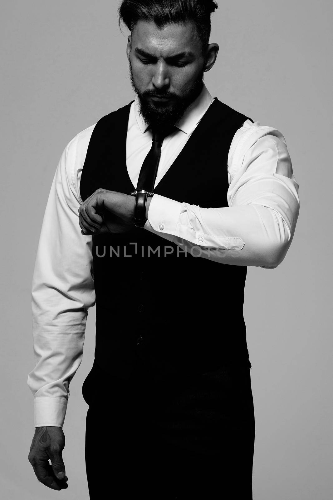 Bearded Hispanic guy in dark vest and white shirt with tie looking at camera with hands in pockets in studio