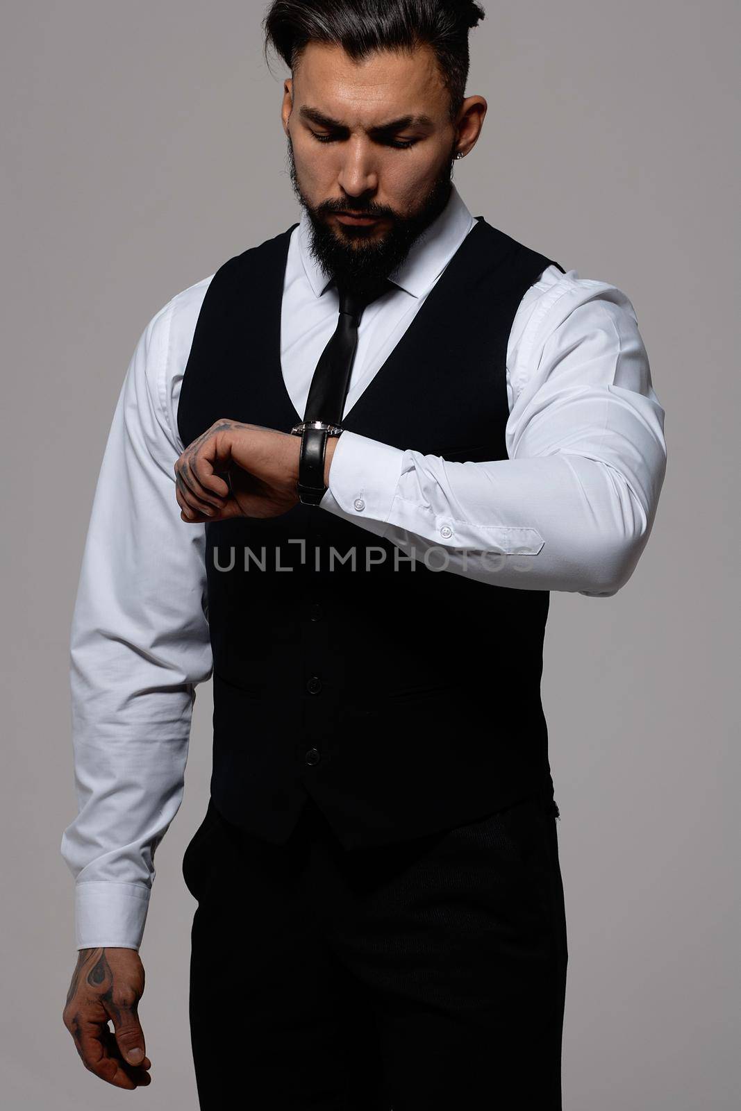 Bearded Hispanic guy in dark vest and white shirt with tie looking at camera with hands in pockets in studio