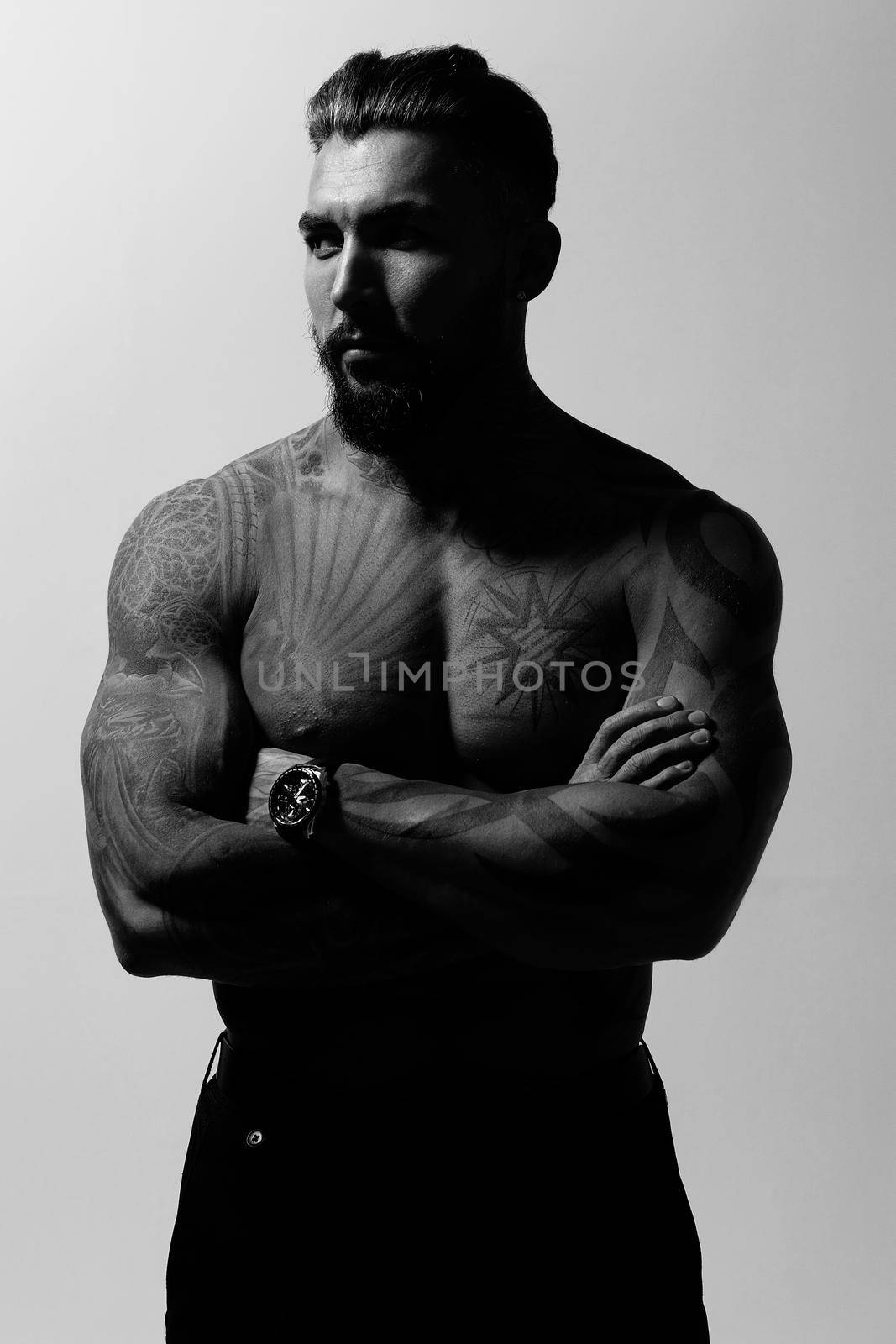 Hispanic shirtless male model with muscular tattooed torso standing with hands in pockets and looking away on gray backdrop