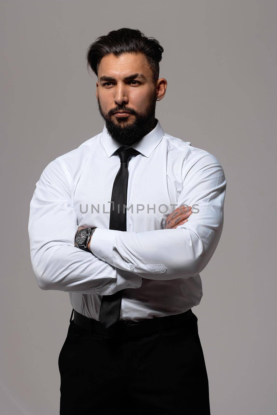 Bearded Hispanic guy in dark vest and white shirt with tie looking at camera with hands in pockets in studio