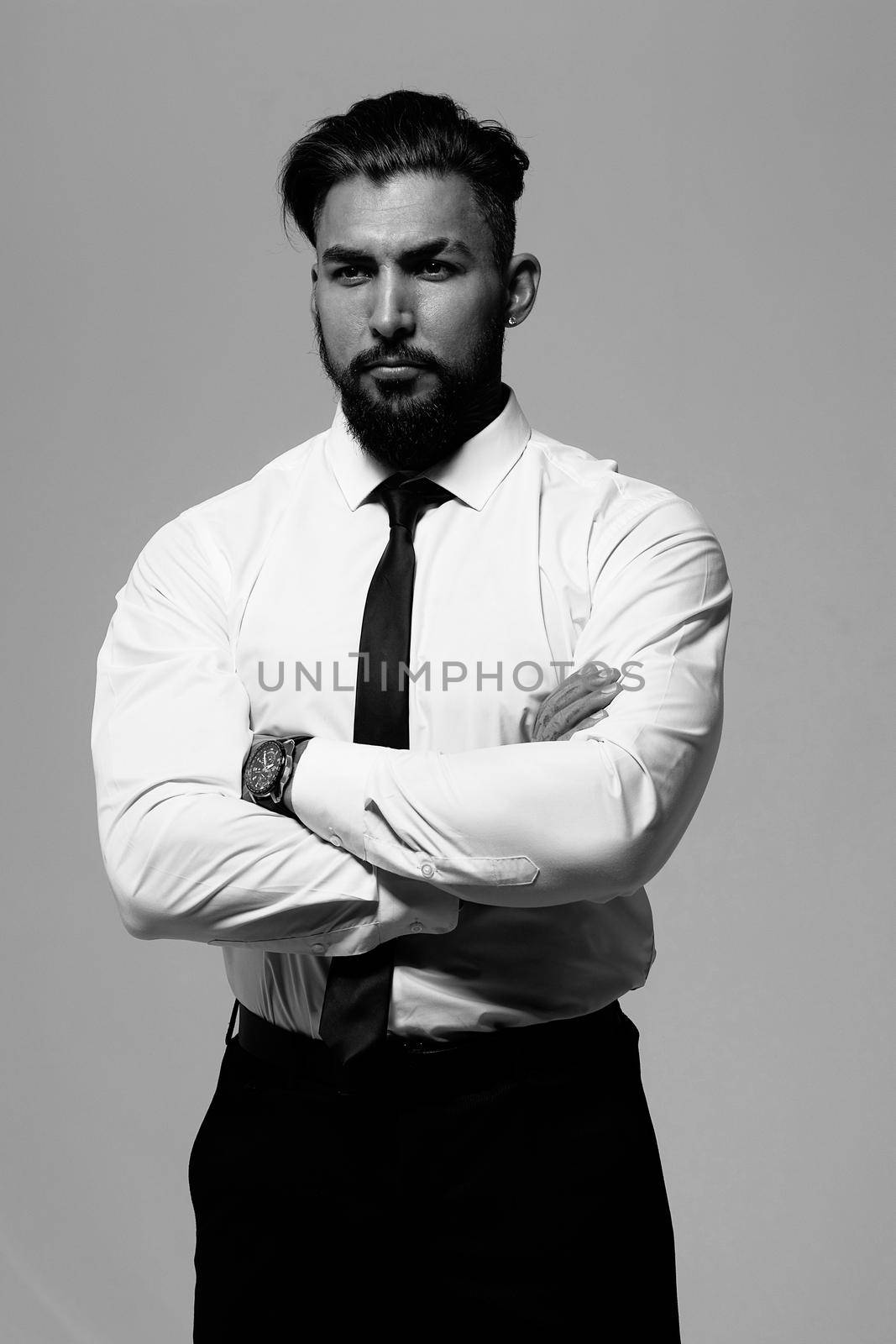 Bearded Hispanic guy in dark vest and white shirt with tie looking at camera with hands in pockets in studio