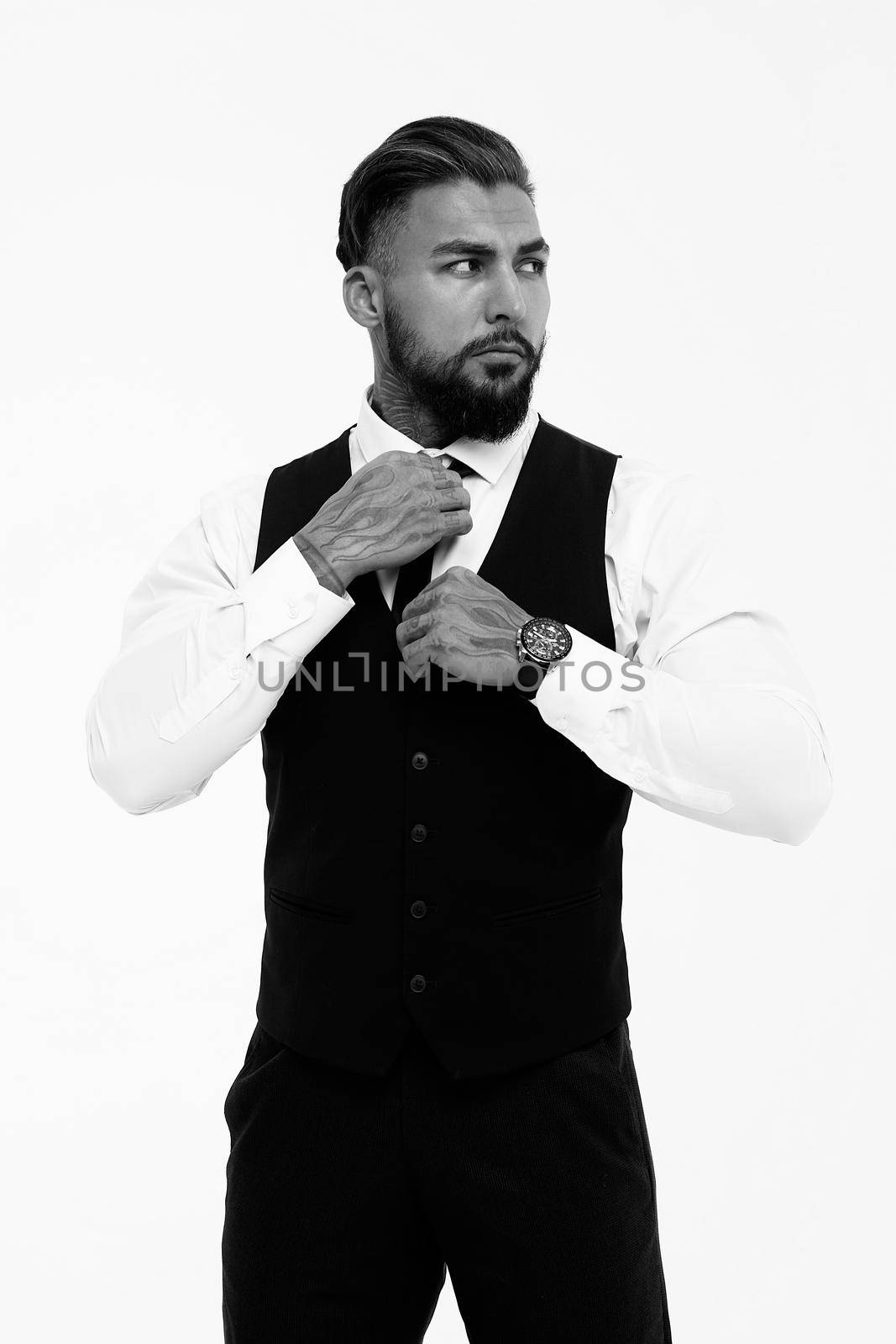 Confident Hispanic bearded man with tattooed hands in white shirt and black trousers adjusting tie and looking away while standing against gray background