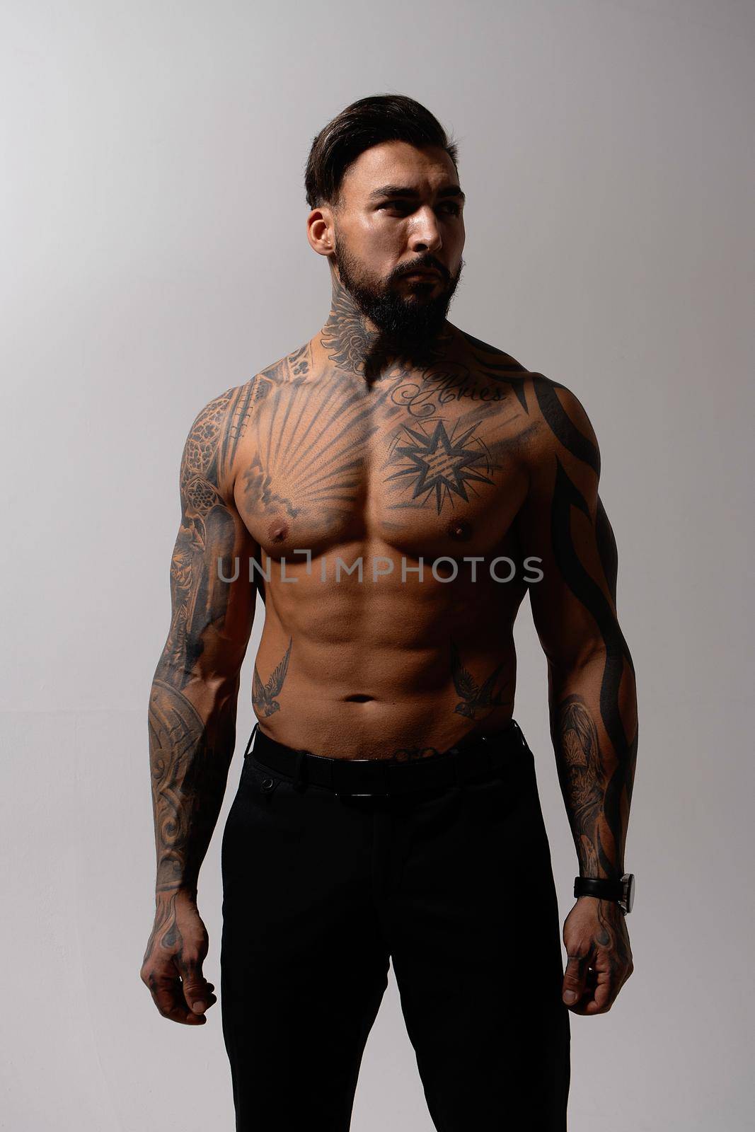 Hispanic shirtless male model with muscular tattooed torso standing with hands in pockets and looking away on gray backdrop