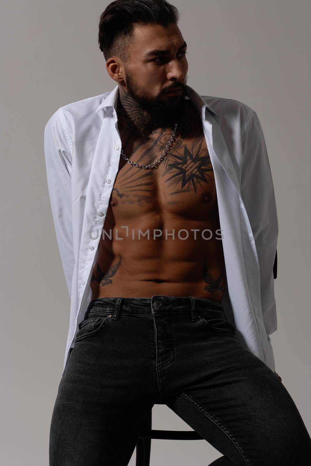 Hispanic shirtless male model with muscular tattooed torso standing with hands in pockets and looking away on gray backdrop