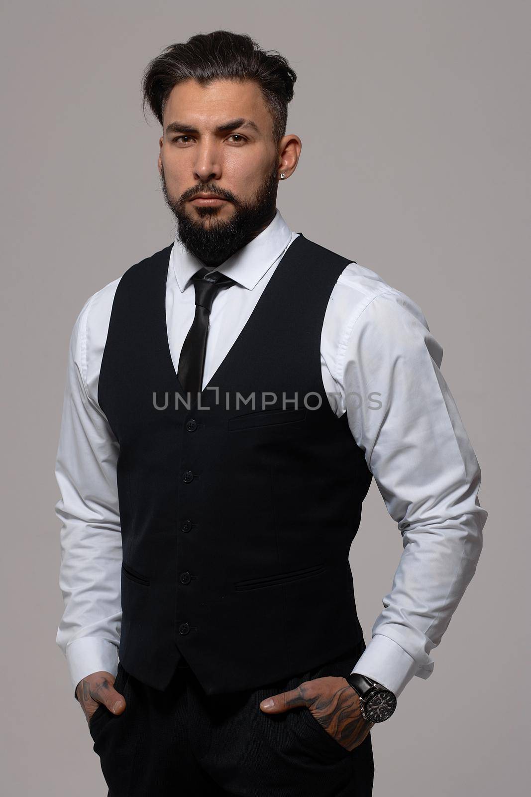 Bearded Hispanic guy in dark vest and white shirt with tie looking at camera with hands in pockets in studio