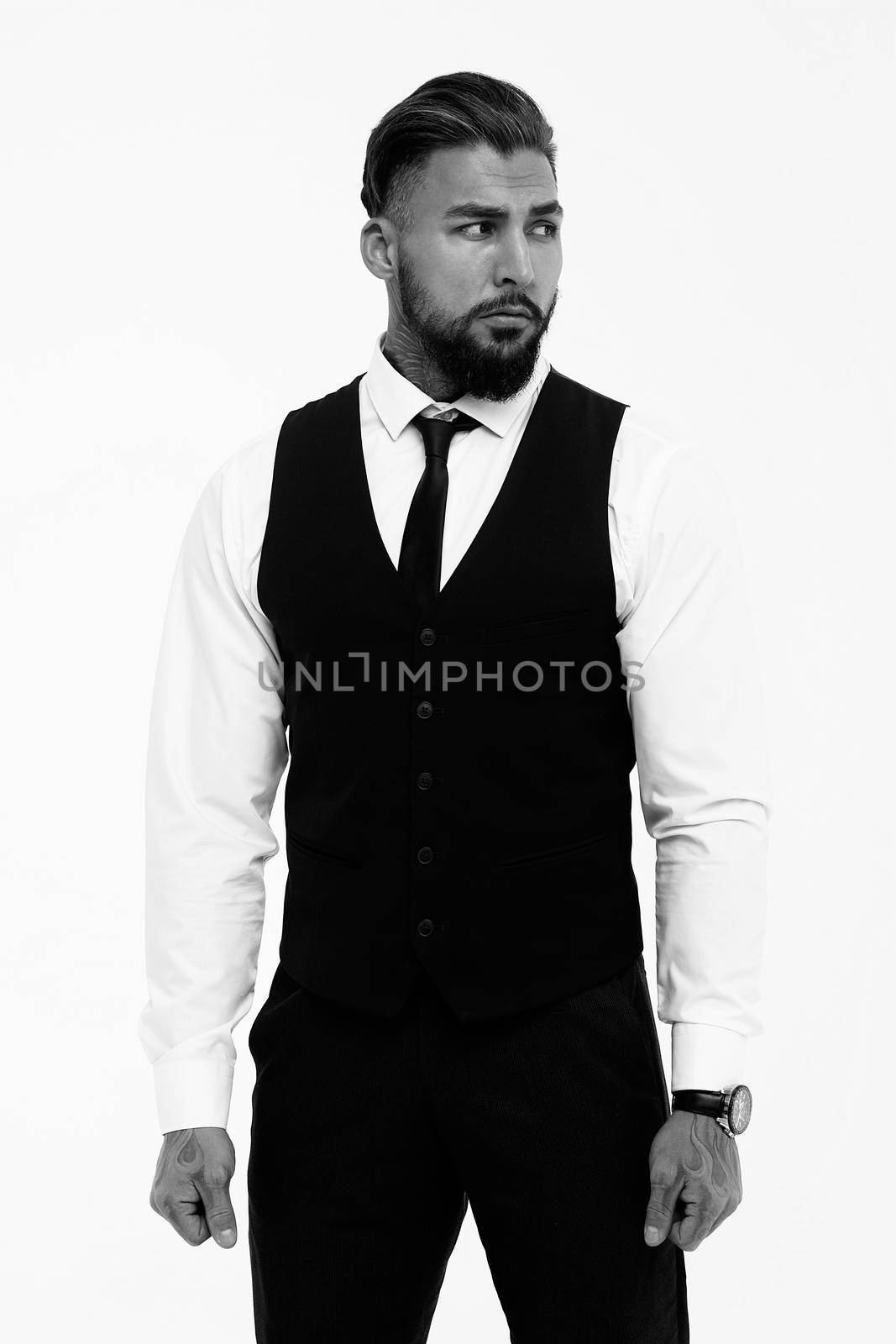 Bearded Hispanic guy in dark vest and white shirt with tie looking at camera with hands in pockets in studio