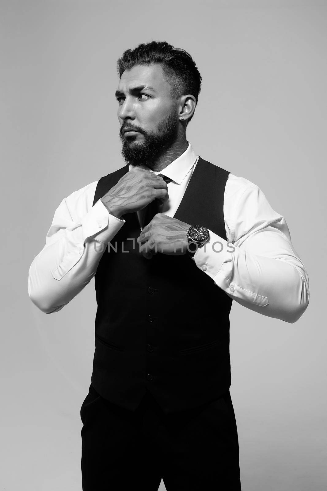 Confident Hispanic bearded man with tattooed hands in white shirt and black trousers adjusting tie and looking away while standing against gray background