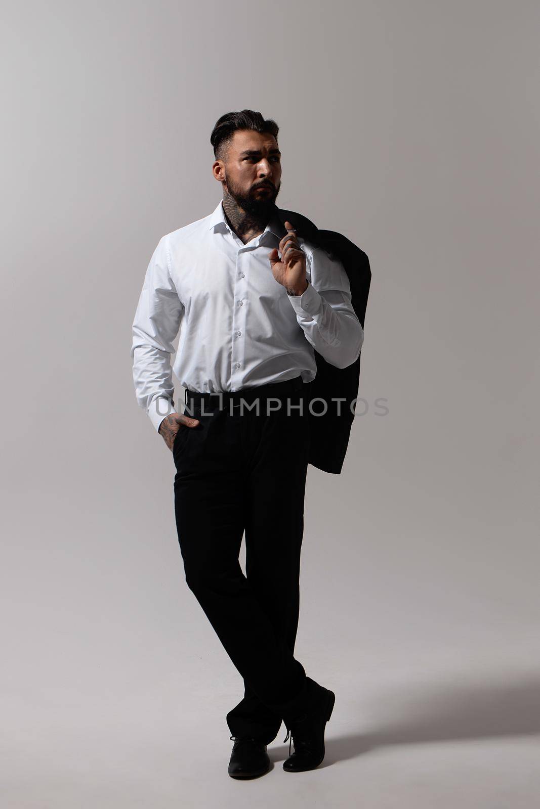 Bearded Hispanic guy in dark vest and white shirt with tie looking at camera with hands in pockets in studio