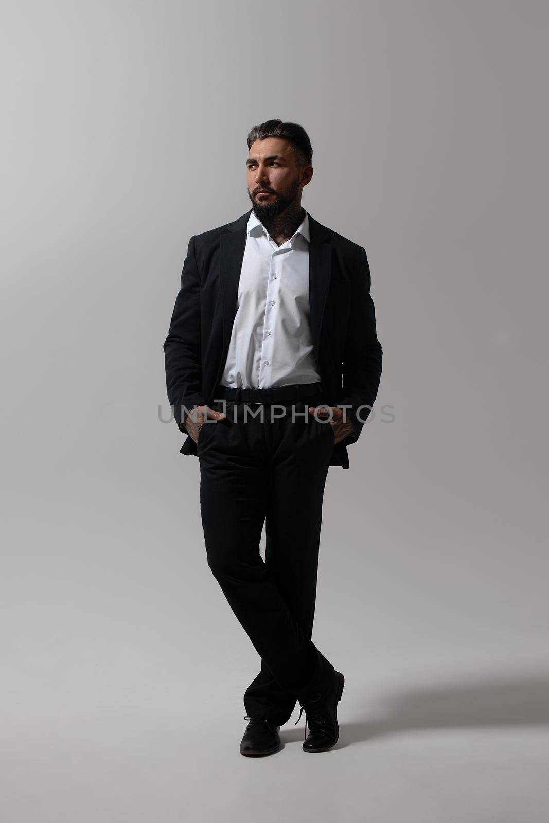 Bearded Hispanic guy in dark vest and white shirt with tie looking at camera with hands in pockets in studio