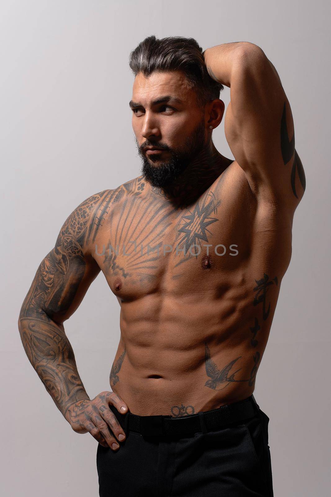 Hispanic shirtless male model with muscular tattooed torso standing with hands in pockets and looking away on gray backdrop