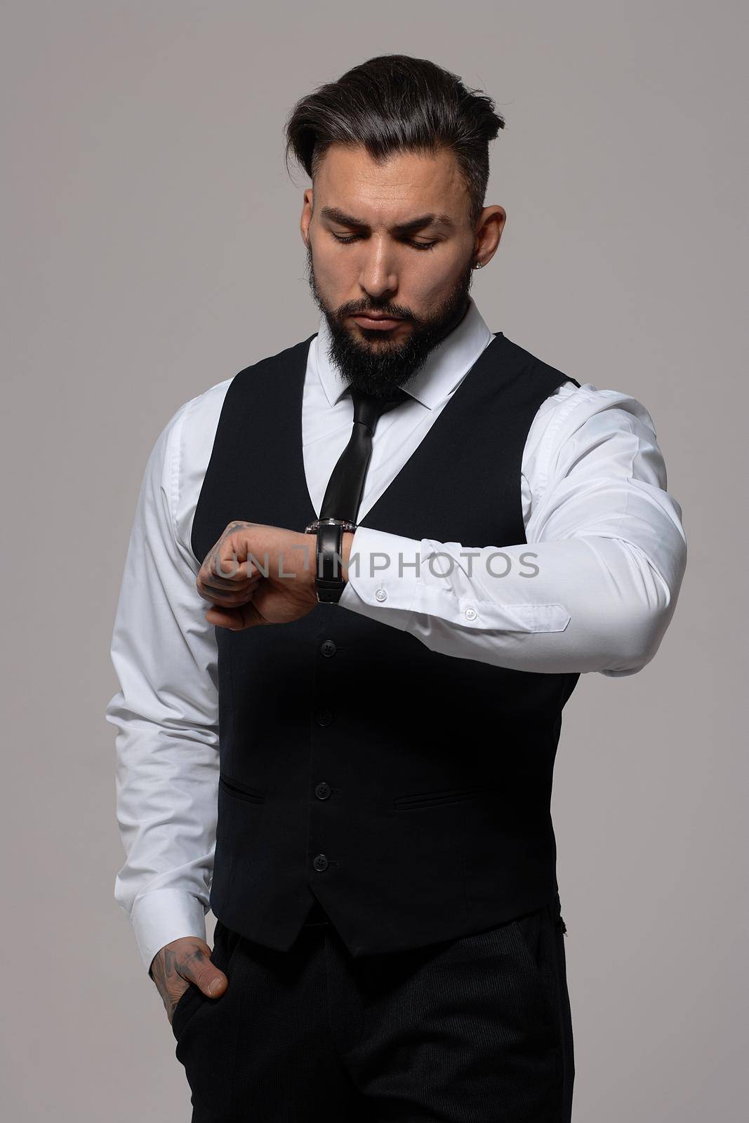 Bearded Hispanic guy in dark vest and white shirt with tie looking at camera with hands in pockets in studio
