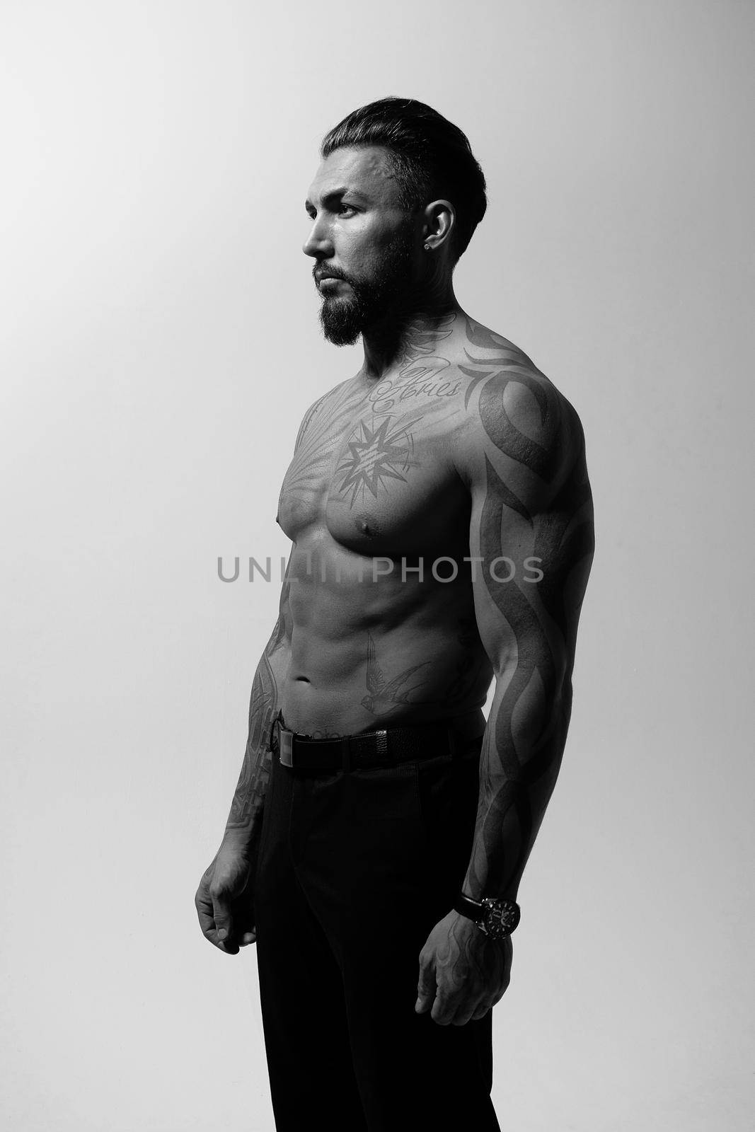 Hispanic shirtless male model with muscular tattooed torso standing with hands in pockets and looking away on gray backdrop