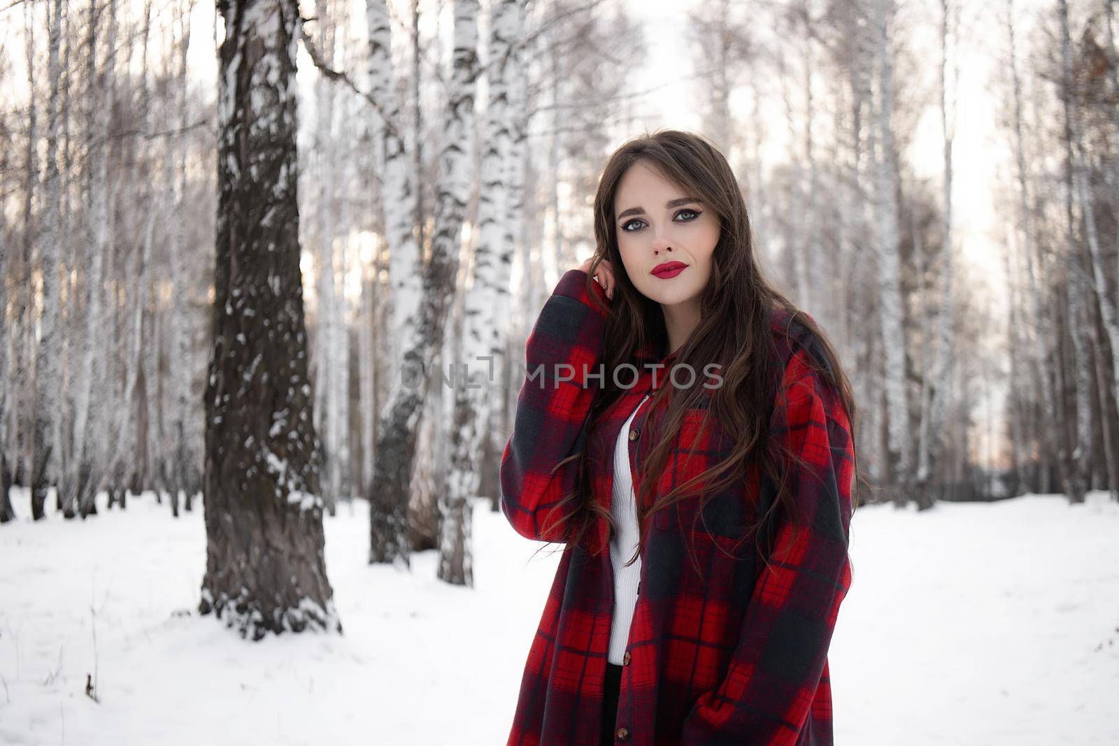 Charming female with red lips and in checkered shirt touching hair while standing in snowy winter woods and looking at camera